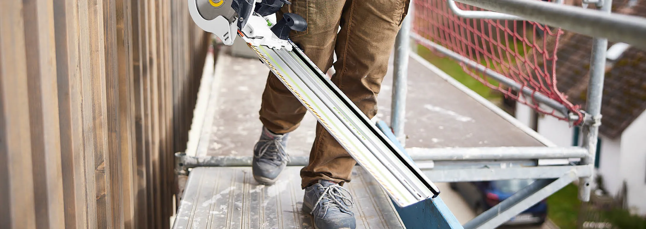 Tradesperson carrying a Festool plunge-cut saw and guide rail across site scaffolding